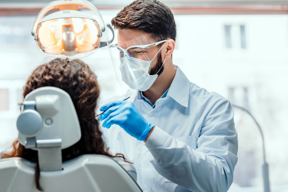 Male Dental Practitioner with brown hair, a mask and clear visor is looking at a female patient who is sitting in a white dental chair.